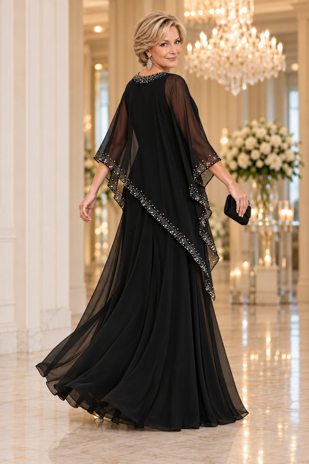 Rear three-quarter view in motion of the black gown, showing the decorated rear neckline, cape drape and floor-length skirt.