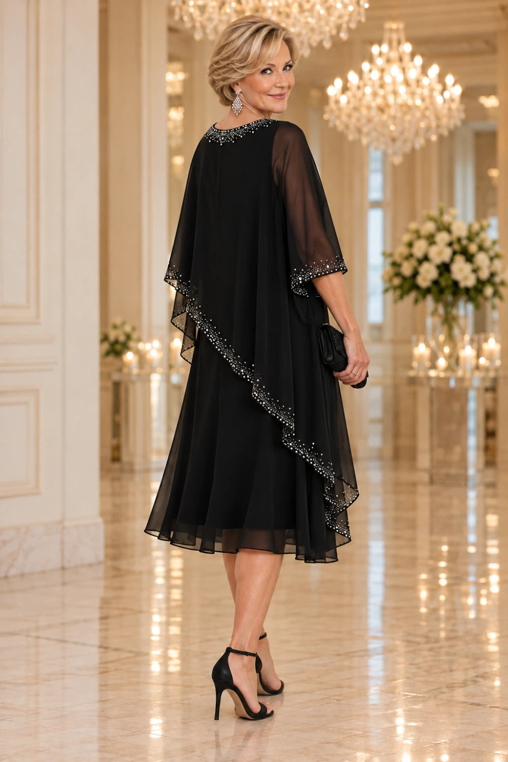 Rear three-quarter view of the black dress with a sheer diagonal cape and decorated round neckline.