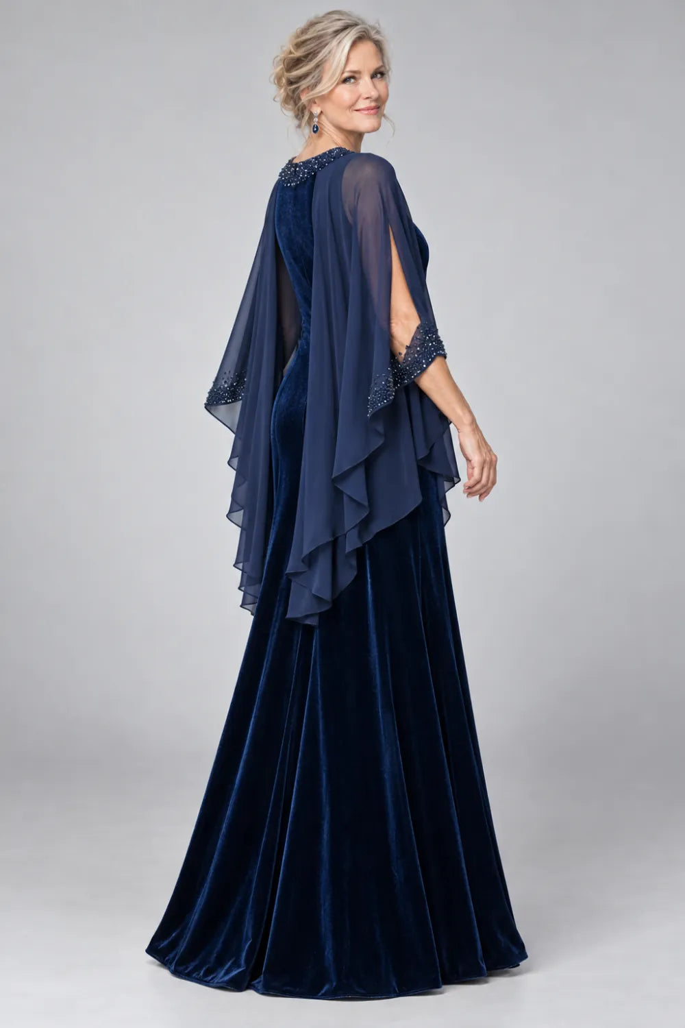 Rear three-quarter view of a navy blue floor-length gown with a sheer cascading cape and decorated arm openings.