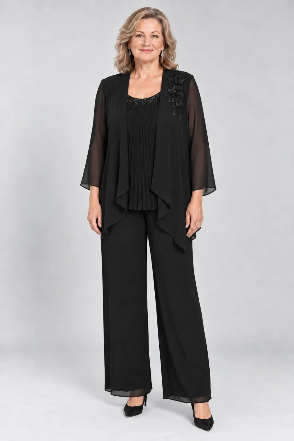 Black pant suit with fine pleats, decorated neckline and floral shoulder motif, front view.